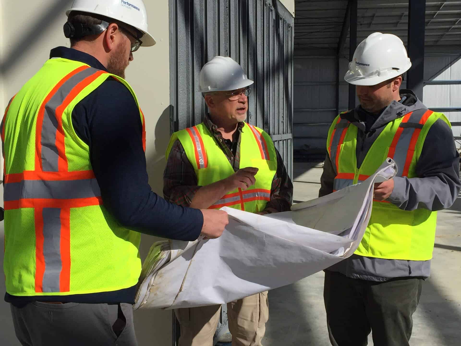 Construction professionals in hard hats huddle around and point at progressive design-build project plans on a jobsite.