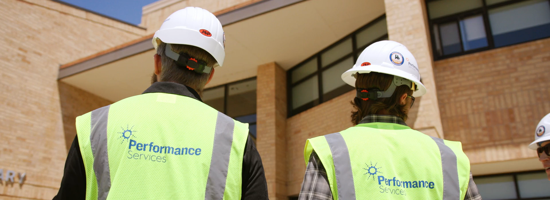 Two Performance Services team members walking into a school building wearing branded safety vests and hard hats